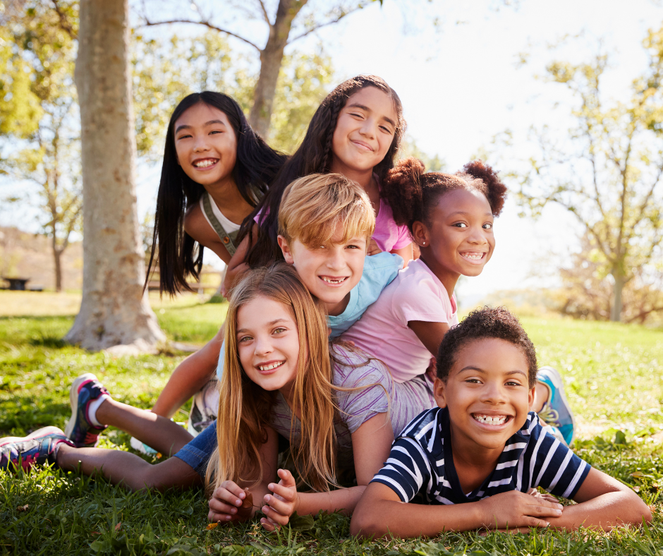 Six smiling kids laying down on the grass on top of each other in a sunny day